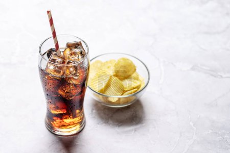 Téléchargez les photos : Verre rafraîchissant de cola avec de la glace, accompagné d'une portion de chips croustillantes. Sur table en pierre avec espace de copie - en image libre de droit