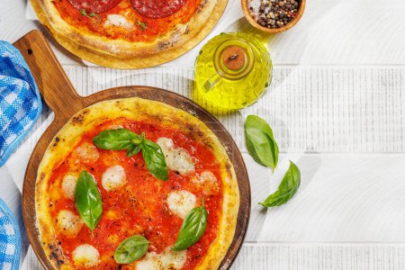 Foto de Pizza casera de margarita y pepperoni, cubierta con tomates frescos, queso mozzarella y hojas aromáticas de albahaca. En la mesa de jardín al aire libre plana con espacio de copia - Imagen libre de derechos