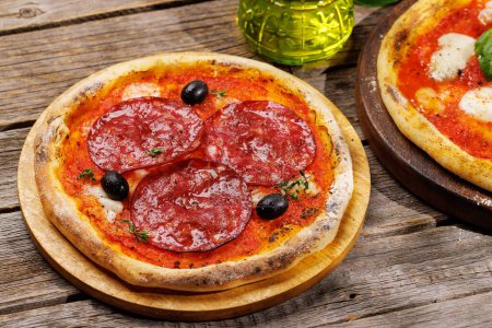 Foto de Pizza casera de margarita y pepperoni, cubierta con tomates frescos, queso mozzarella y hojas aromáticas de albahaca. En la mesa de jardín al aire libre - Imagen libre de derechos