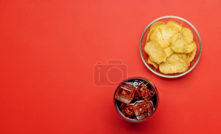 Téléchargez les photos : Refreshing glass of cola with ice, accompanied by a serving of crispy chips. Over red background with copy space. Flat lay - en image libre de droit