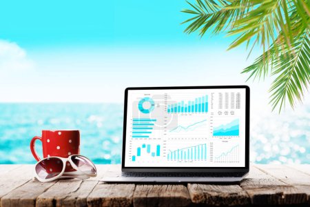 Photo for Laptop business reports and charts and coffee cup on wooden table in front of sunny sea and palm leaves. Work and travel or remote business concept - Royalty Free Image