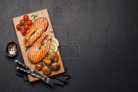 Téléchargez les photos : Des steaks de saumon grillés et des pommes de terre sur une planche de bois, un délice appétissant. Pose plate avec espace de copie - en image libre de droit