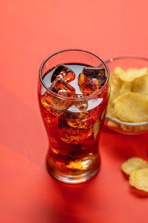 Téléchargez les photos : Verre rafraîchissant de cola avec de la glace, accompagné d'une portion de chips croustillantes. Sur fond rouge - en image libre de droit