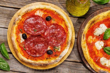 Foto de Pizza casera de margarita y pepperoni, cubierta con tomates frescos, queso mozzarella y hojas aromáticas de albahaca. En la mesa de jardín al aire libre plana - Imagen libre de derechos