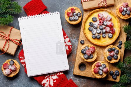 Foto de Delicia festiva: cupcakes de Navidad adornados con bayas. Puesta plana con bloc de notas para su receta o texto de saludo - Imagen libre de derechos