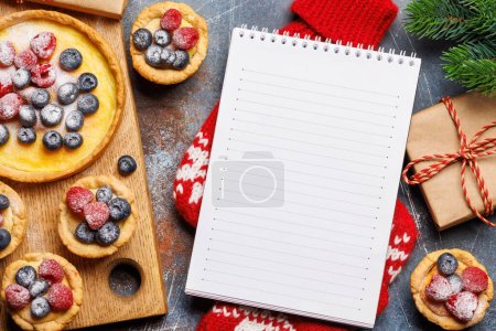 Foto de Delicia festiva: cupcakes de Navidad adornados con bayas. Puesta plana con bloc de notas para su receta o texto de saludo - Imagen libre de derechos