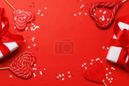 Téléchargez les photos : Sucettes de coeur : friandises sucrées et boîtes-cadeaux sur fond rouge avec espace texte. Pose plate carte Saint Valentin - en image libre de droit