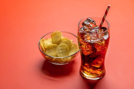 Verre rafraîchissant de cola avec de la glace, accompagné d'une portion de chips croustillantes. Sur fond rouge avec espace de copie