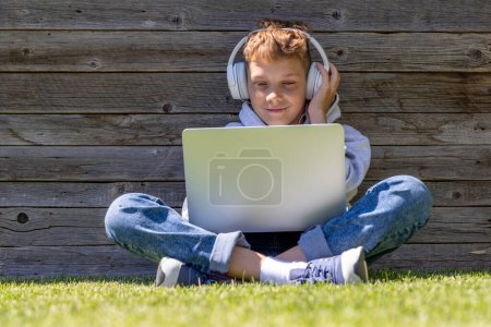 Téléchargez les photos : Un garçon utilisant un ordinateur portable et un casque à l'extérieur. Concept d'éducation ou de divertissement - en image libre de droit