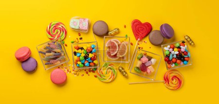 Various colorful candies, lollipops, and macaroons. Flat lay sweets over yellow background