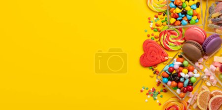 Various colorful candies, lollipops, and macaroons. Flat lay sweets over yellow background with copy space