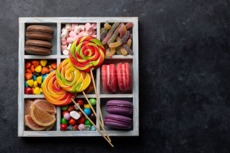 Various colorful candies, lollipops, and macaroons. Flat lay sweets in box over stone background with copy space