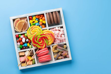 Various colorful candies, lollipops, and macaroons. Flat lay sweets in box over blue background with copy space