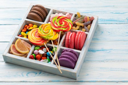 Foto de Varios caramelos coloridos, piruletas y macarrones. Dulces en caja sobre fondo de madera con espacio para copiar - Imagen libre de derechos
