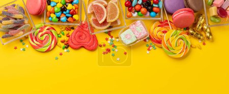 Various colorful candies, lollipops, and macaroons. Flat lay over yellow background with copy space