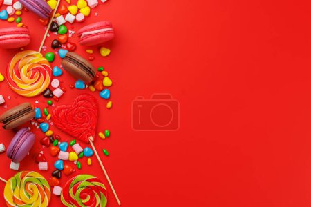 Téléchargez les photos : Divers bonbons colorés, sucettes et macarons. Pose plate sur fond rouge avec espace de copie - en image libre de droit
