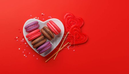 Téléchargez les photos : Divers macarons colorés dans un bol en forme de coeur. Amour bonbons sur fond rouge avec espace de copie - en image libre de droit