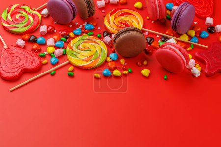 Divers bonbons colorés, sucettes et macarons. Sur fond rouge avec espace de copie