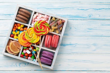 Téléchargez les photos : Divers bonbons colorés, sucettes et macarons. Poser des bonbons plats en boîte sur un fond en bois avec espace de copie - en image libre de droit