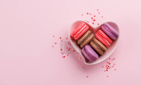Various colorful macaroons in heart shaped bowl. Love sweets over pink background with copy space