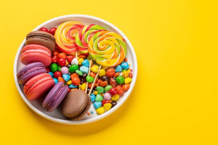 Varios caramelos coloridos, piruletas y macarrones. Sobre fondo amarillo con espacio de copia