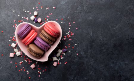 Various colorful macaroons in heart shaped bowl. Love sweets over stone background with copy space