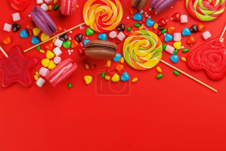 Divers bonbons colorés, sucettes et macarons. Pose plate sur fond rouge avec espace de copie
