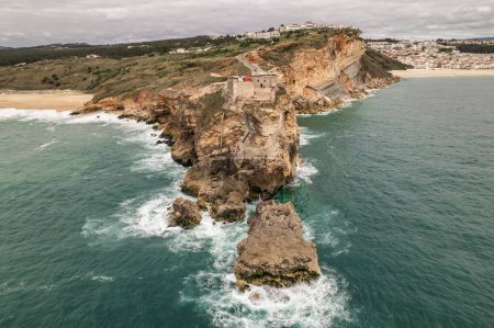 Photo for Aerial view of an old lighthouse on a cliff with a fortress on the coast of the Atlantic ocean in Nazare town, Portugal. Flying towards the lighthouse near the Nazare city in Portugal. - Royalty Free Image
