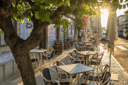 Photo for Outdoor cafe on a beautiful street with traditional azulejos Portuguese houses in Tavira town at sunset, Algarve region, Portugal. - Royalty Free Image