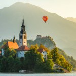 Lake Bled, Slovenia, in summer. A close-up view of the Pilgrimage Church on a small island, Bled Castle, and a hot air balloon at sunrise over the picturesque Bled Lake.
