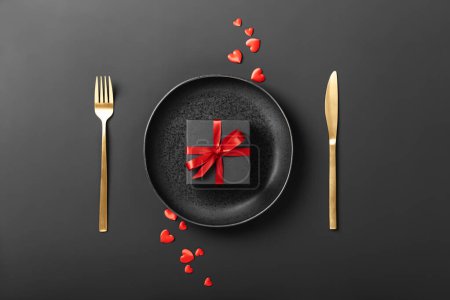 Foto de Beautiful romantic table setting with plate, fork, knife and gift box on dark background - Imagen libre de derechos
