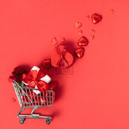 Foto de Shopping trolley with gift boxes, roses, chocolate and red hearts on red background. Online stores. Winter Holiday Sales, Seasonal Sales, Black Friday, February 14 Valentines Day. Discounts and on - Imagen libre de derechos
