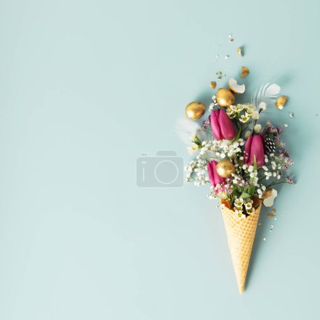 Photo for Easter composition. Ice cream cone with beautiful flowers and golden easter eggs on blue background flat lay - Royalty Free Image