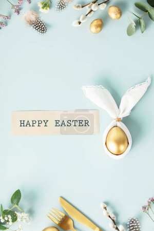 Foto de Stylish Easter flat lay with golden egg in easter bunny napkin, golden quail eggs, feathers, cutlery and spring flowers on blue background. Minimalist modern Easter table decorations. Top view flat - Imagen libre de derechos