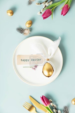 Téléchargez les photos : Élégant plat de Pâques avec oeuf doré dans une serviette de lapin de Pâques sur assiette blanche, œufs de caille dorée, plumes, couverts et fleurs de printemps sur fond bleu. Décorations de table de Pâques modernes minimalistes - en image libre de droit
