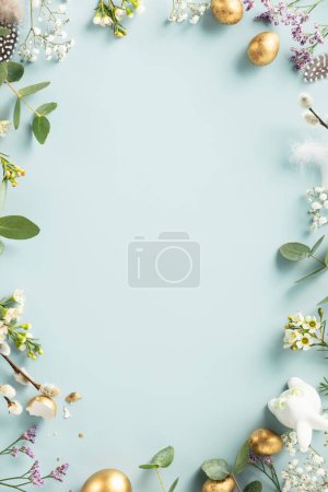 Foto de Easter table decorations. Happy Easter concept with golden easter eggs, feathers and spring flowers on blue background. Flat lay top view copy space - Imagen libre de derechos
