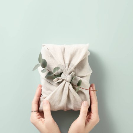 Téléchargez les photos : Furoshiki tissu emballage de cadeaux. Main féminine tenant un cadeau dans un emballage en tissu réutilisable respectueux de l'environnement. Petite entreprise, idée de shopping éthique. Présente emballée sans plastique. Style de vie zéro déchet - en image libre de droit