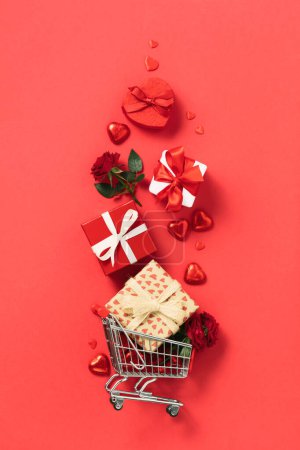 Téléchargez les photos : Shopping trolley with gift boxes, roses, chocolate and red hearts on red background. Online stores. Winter Holiday Sales, Seasonal Sales, Black Friday, February 14 Valentines Day. Discounts and on - en image libre de droit