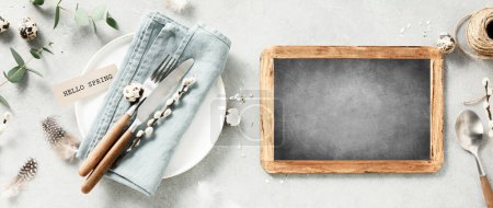 Photo for Banner. Table setting. A fashionable minimalistic plate with a linen napkin, knife, fork, chalkboard, Easter eggs and feathers on a gray background. Top view. Happy Easter holiday concept for cafes - Royalty Free Image