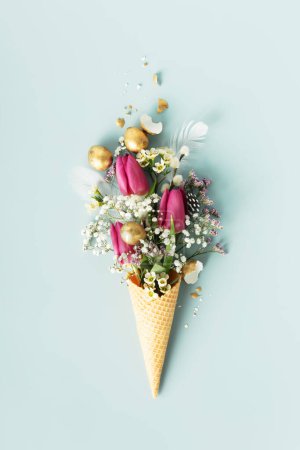 Photo for Easter composition. Ice cream cone with beautiful flowers and golden easter eggs on blue background flat lay - Royalty Free Image
