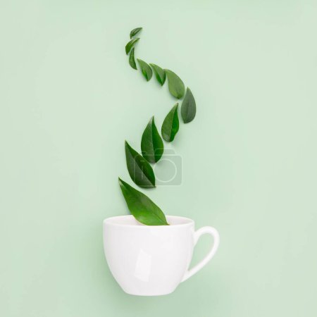 Téléchargez les photos : Une tasse de thé blanc avec un sillage fantaisiste de feuilles vertes suggérant un mélange de thé naturel et aromatique, sur un fond vert doux. - en image libre de droit