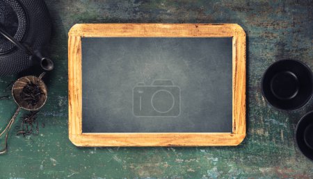 Photo for A rustic tea time setting featuring a cast iron teapot, cups, and a loose tea strainer, next to a blank chalkboard on a worn wooden surface. - Royalty Free Image