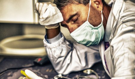 Téléchargez les photos : Médecin stressé dans le masque de weraing pandémie covid et gants tenant smartphone devant l'écran d'ordinateur. - en image libre de droit