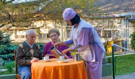 Foto de Joven asiática camarera sirviendo desayuno a ancianos jubilados pareja en un hospital rehabilitación jardín. - Imagen libre de derechos