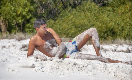 Téléchargez les photos : Portrait d'un homme caucasien heureux se relaxant sur une belle plage couverte de sable sur le corps. - en image libre de droit