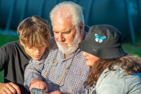 Téléchargez les photos : Grand-père examinant les images d'un appareil photo compact en plein air avec ses petits-enfants - en image libre de droit