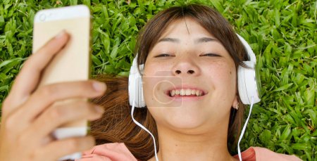 Téléchargez les photos : Heureuse adolescente asiatique allongée sur l'herbe entendre de la musique. Concept de légèreté - en image libre de droit
