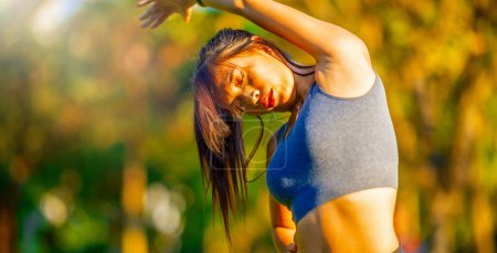 Photo for Beautiful asian girl dressed in sporty clothes stretching her body in a city park. - Royalty Free Image
