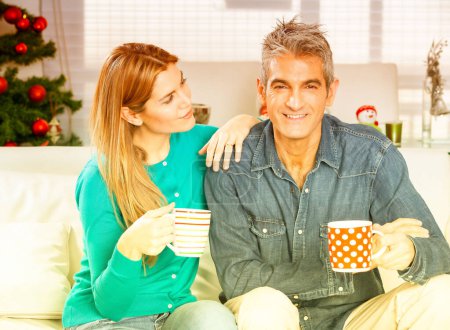 Foto de Feliz joven pareja sonriente bebiendo café sentado en el sofá de casa para Navidad - Imagen libre de derechos