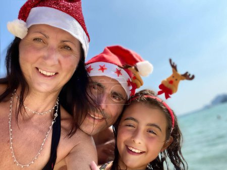 Foto de Navidad tropical familiar. Chica joven junto con sus padres con sombreros de Navidad en una hermosa playa. - Imagen libre de derechos